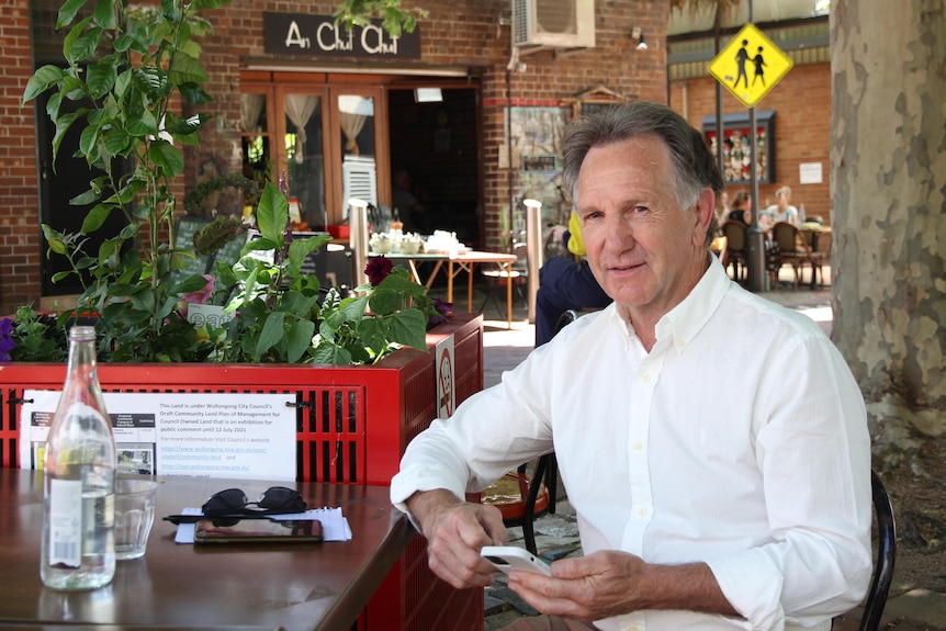 a man in a white shirt sits at a cafe table under a tree