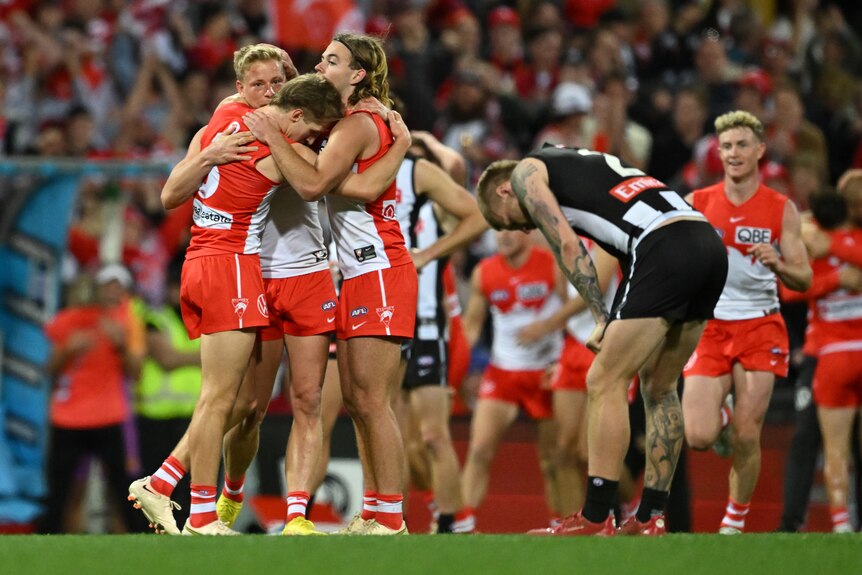 A group of victorious Sydney Swans AFL players hug each other in celebration as a dejected Collingwood player bends over.