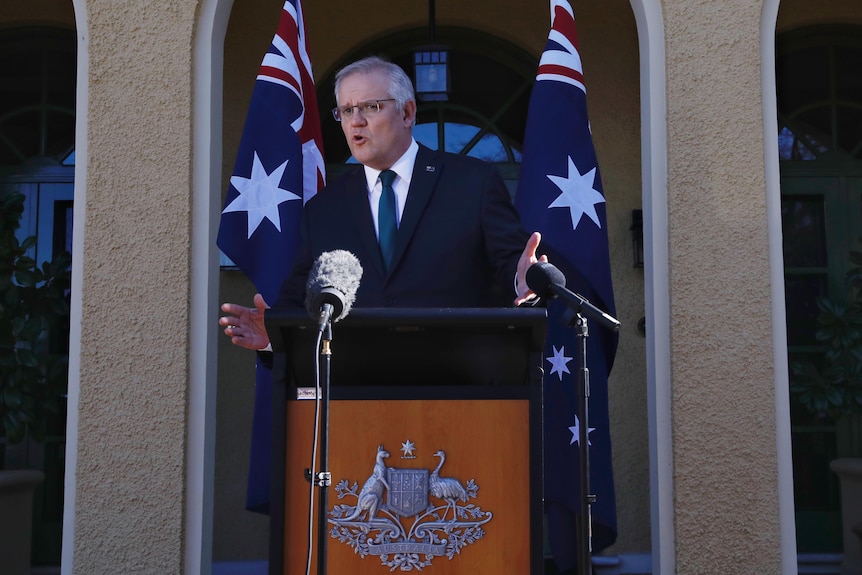 The Prime Minister stands before a lectern and flags in the courtyard of the Lodge.