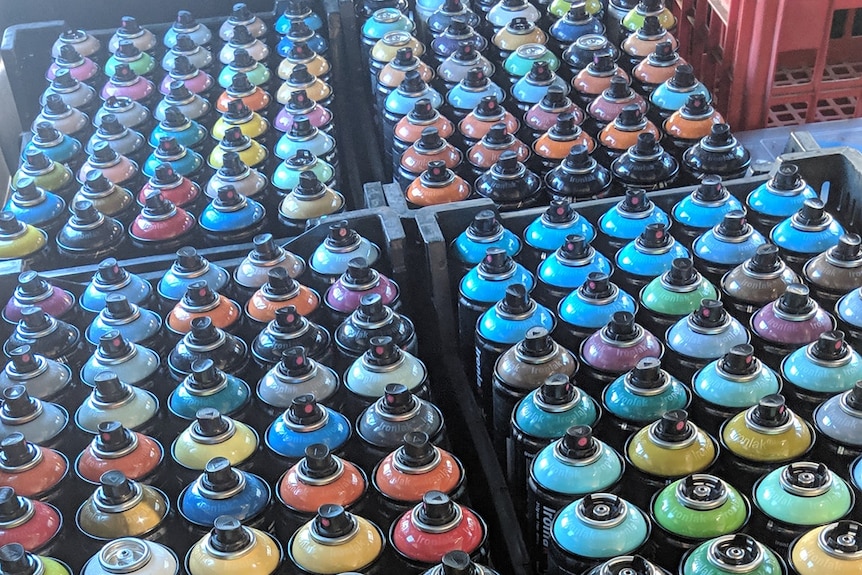 Four black crates of spray paint cans sit on carpet.