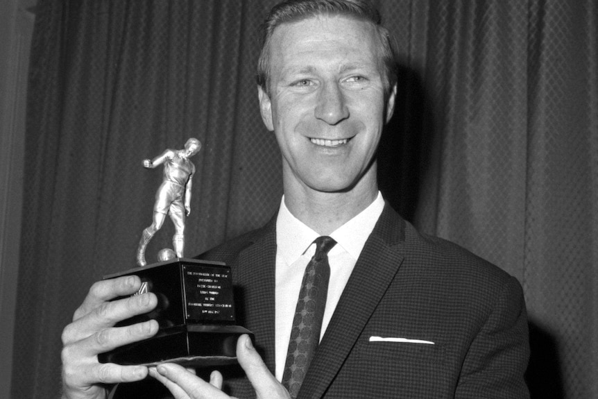 A black and white photo of Jack Charlton, who is wearing a nice suit, smiling while holding a trophy