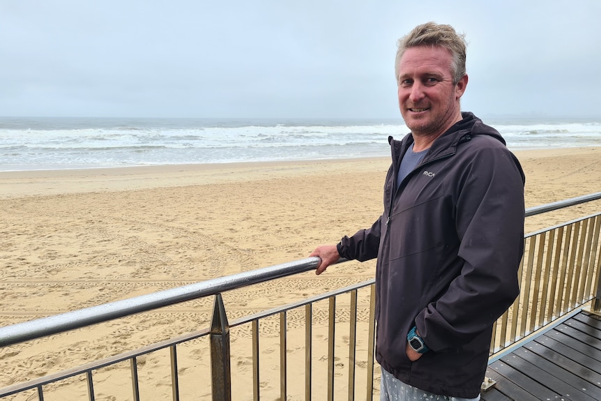 Grant Thomas stands in a wind jacket holding a railing by the beach.