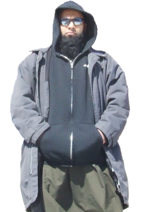 A man with a beard wears a hoodie, black boots and reflective sunglasses. 