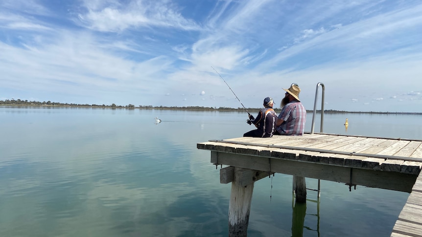 A man and boy sitting on the end of a jetty at a lake with fishing rods, blue sky with white clouds reflected in water.