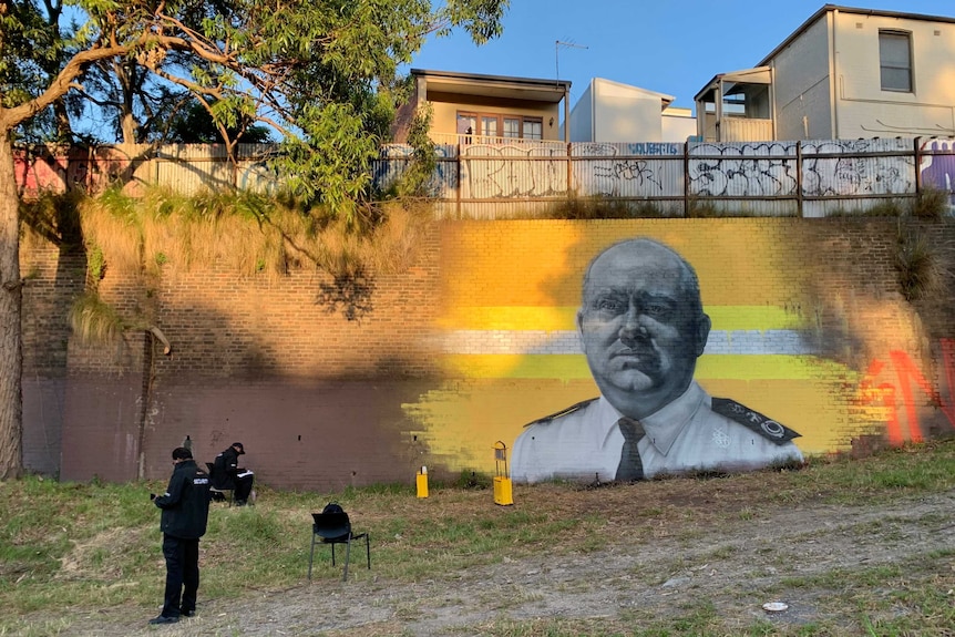 Security guards sit on the grass in front of a wall mural of Shane Fitzsimmons