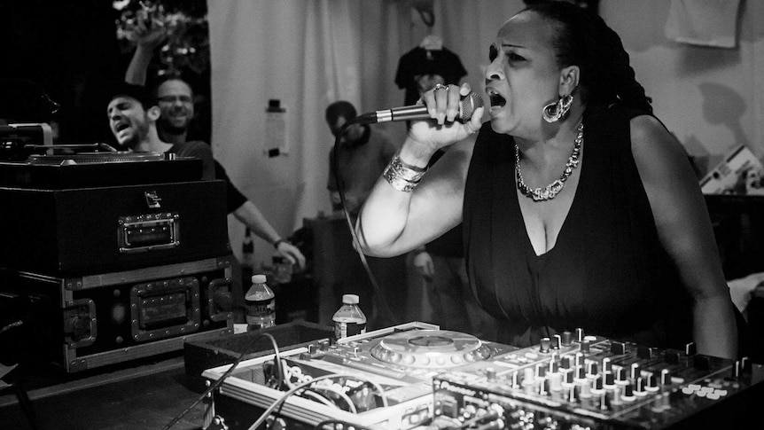 A Jamaican woman wearing jewels and a black shirt singing into a microphone in front of a DJ turntable