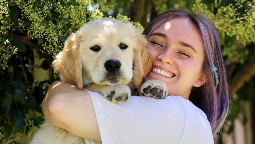 A woman with puruple hair smiles with her eyes shut while holding a golden haired puppy up in their arms.