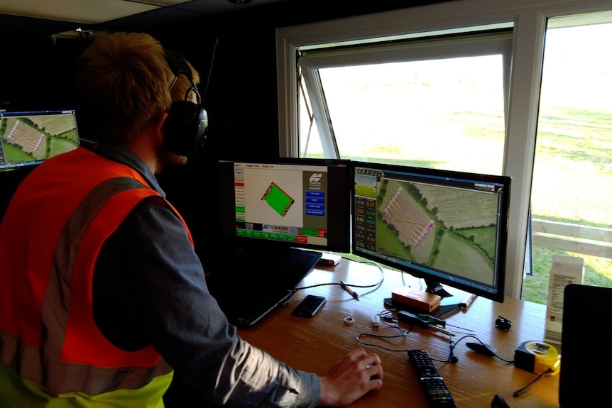 A man wearing earmuffs sitting in a control room looking a computer monitors, with windows and a paddock in the background.