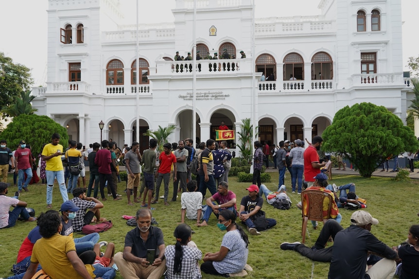 Groups of people sit and stand on the grass outside an ornate, white, colonial-era building. Guards stand on a balcony