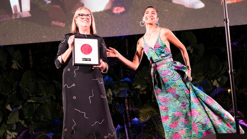 Two women stand on a stage.  One is holding a framed award. 