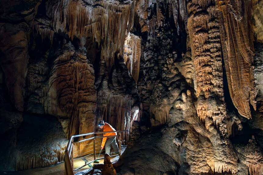 A woman in a fluro orange jacket and headlamp sweeps the path beneath vast and intricate limestone formations inside a cave.