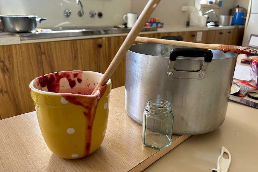 A half-filled jug of homemade raspberry jam sits on a bench next to a large pot and several wooden spoons.