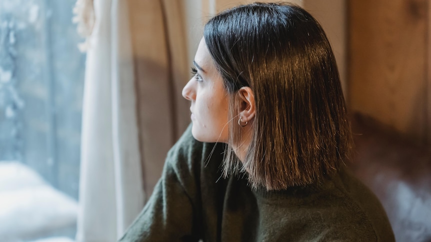 A woman with short dark hair wearing a khaki green long sleeved shirt sits on a windowsill looking out a window.