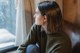 A woman with short dark hair wearing a khaki green long sleeved shirt sits on a windowsill looking out a window.
