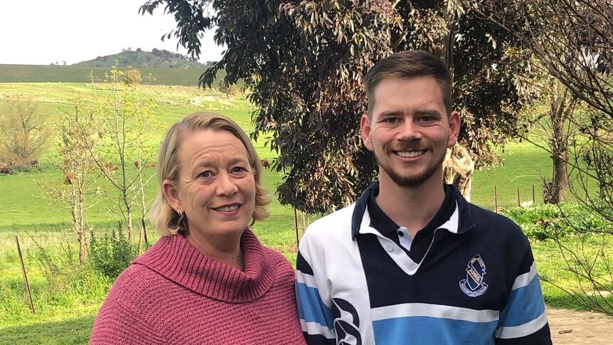 Middle-aged woman with blonde hair and wearing a pink knitted jumper with a taller high school student in a blue school jersey.