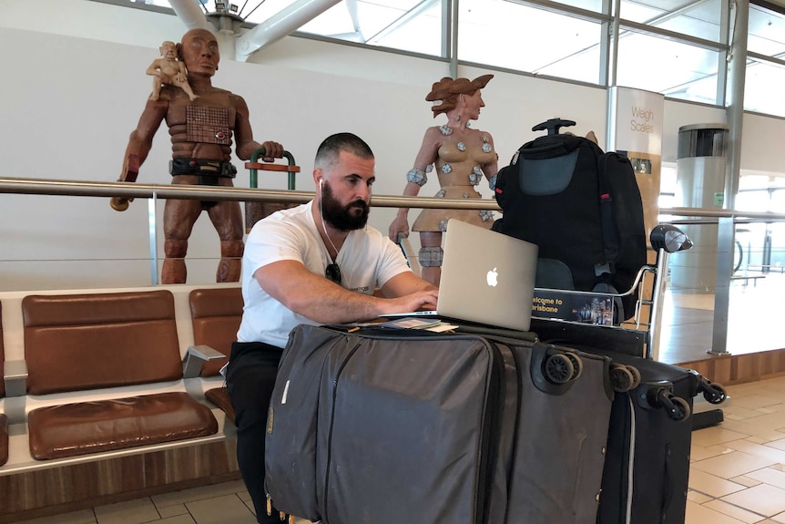 A man types on a laptop on top of his luggage in an airport terminal.