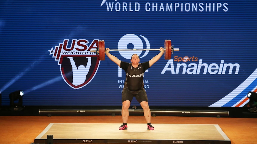 A wide view shows Laurel Hubbard holding the bar and weights above her head during a weightlifting competition.