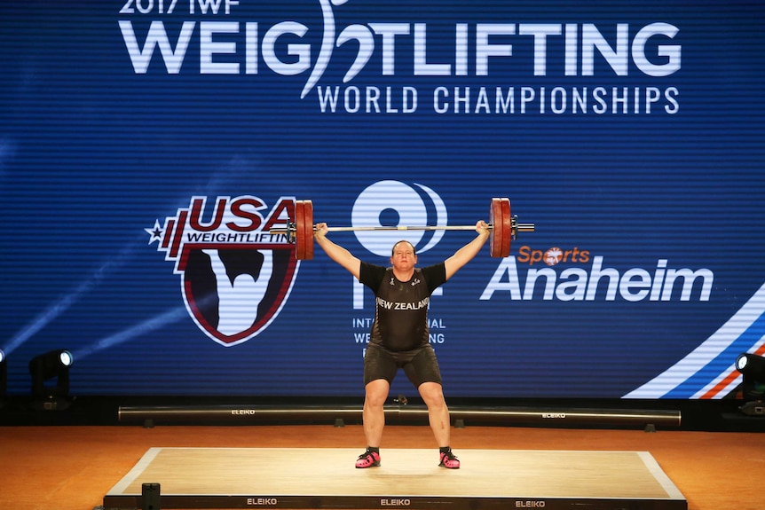 A wide view shows Laurel Hubbard holding the bar and weights above her head during a weightlifting competition.