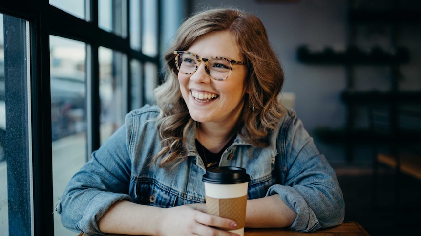 A woman sits at a table in a cafe with a coffee and smiling.