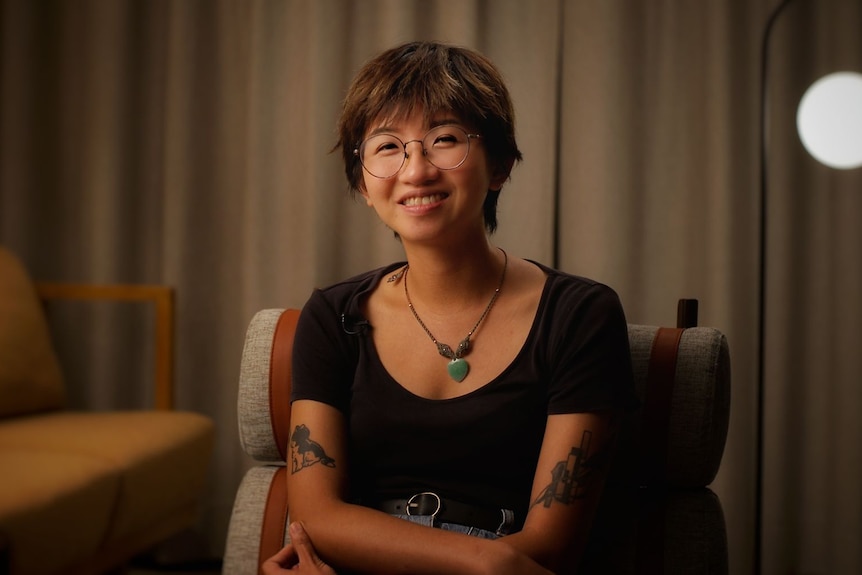 A young woman with short hair, tattoos and wire-rimmed glasses smiles while folding her arms