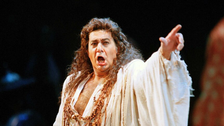 Placido Domingo performs on stage with long curly hair and an open white shirt.