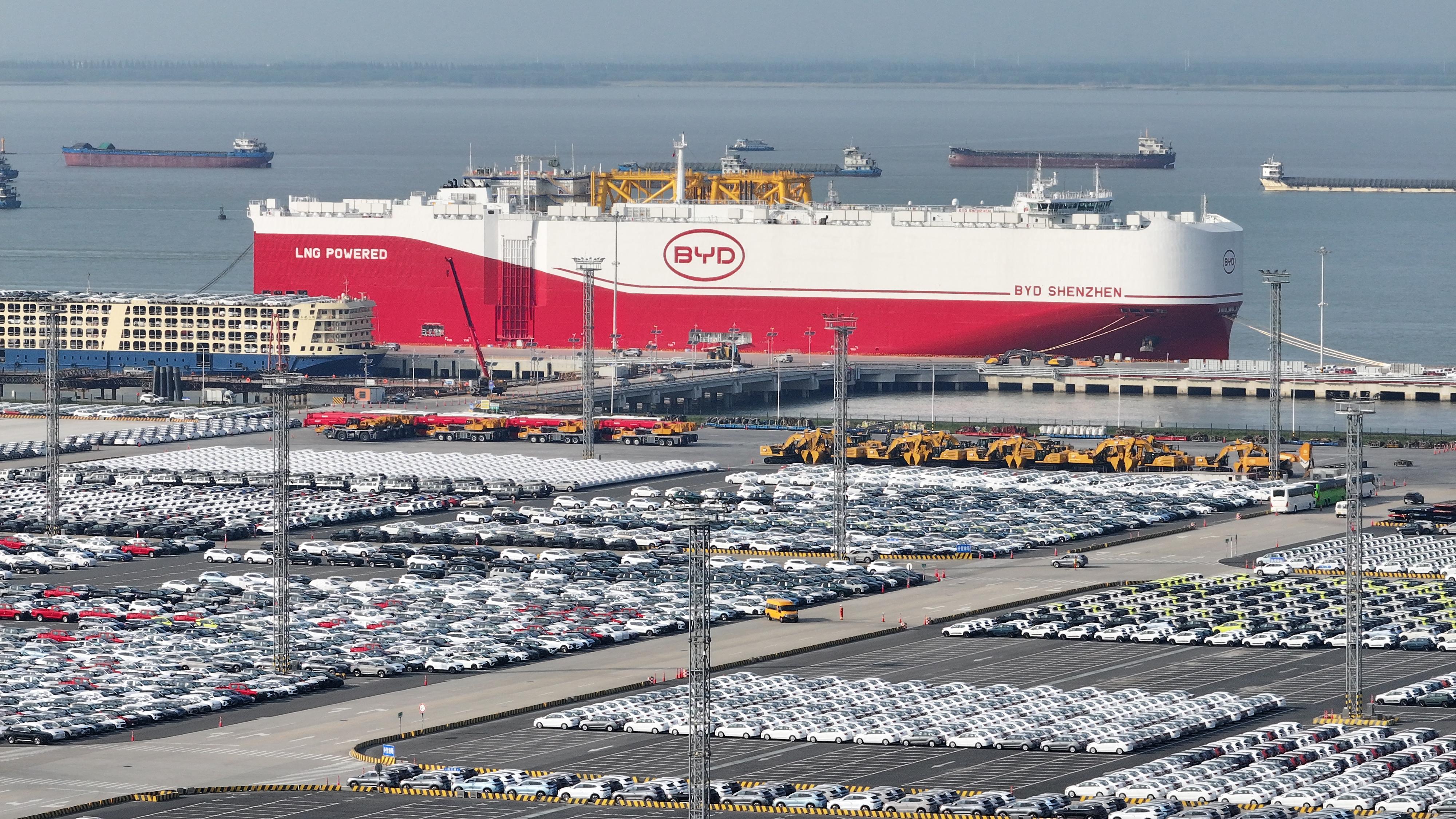 Rows of vehicles lined up at a port near a large white and red ship with a logo saying "BYD".