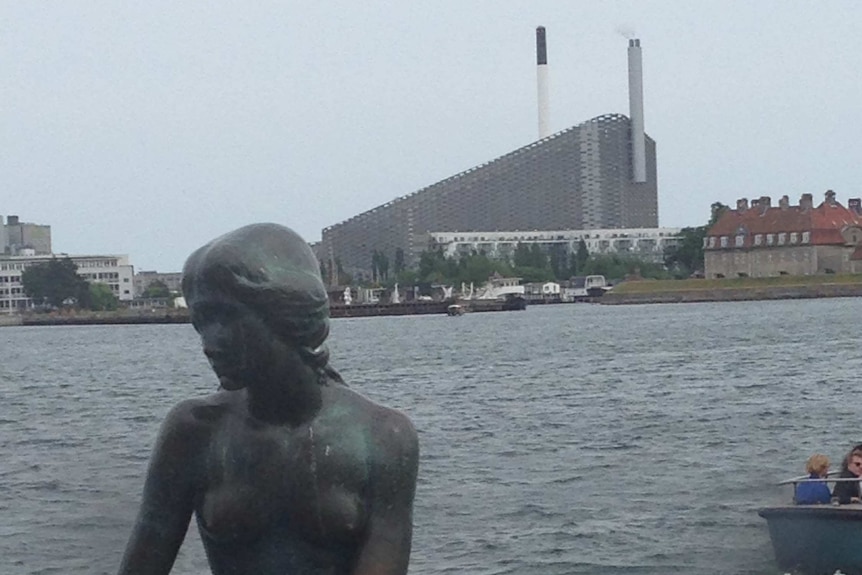 View of the Little Mermaid statue by the water, with a large, sloped building with two large chimneys in the background