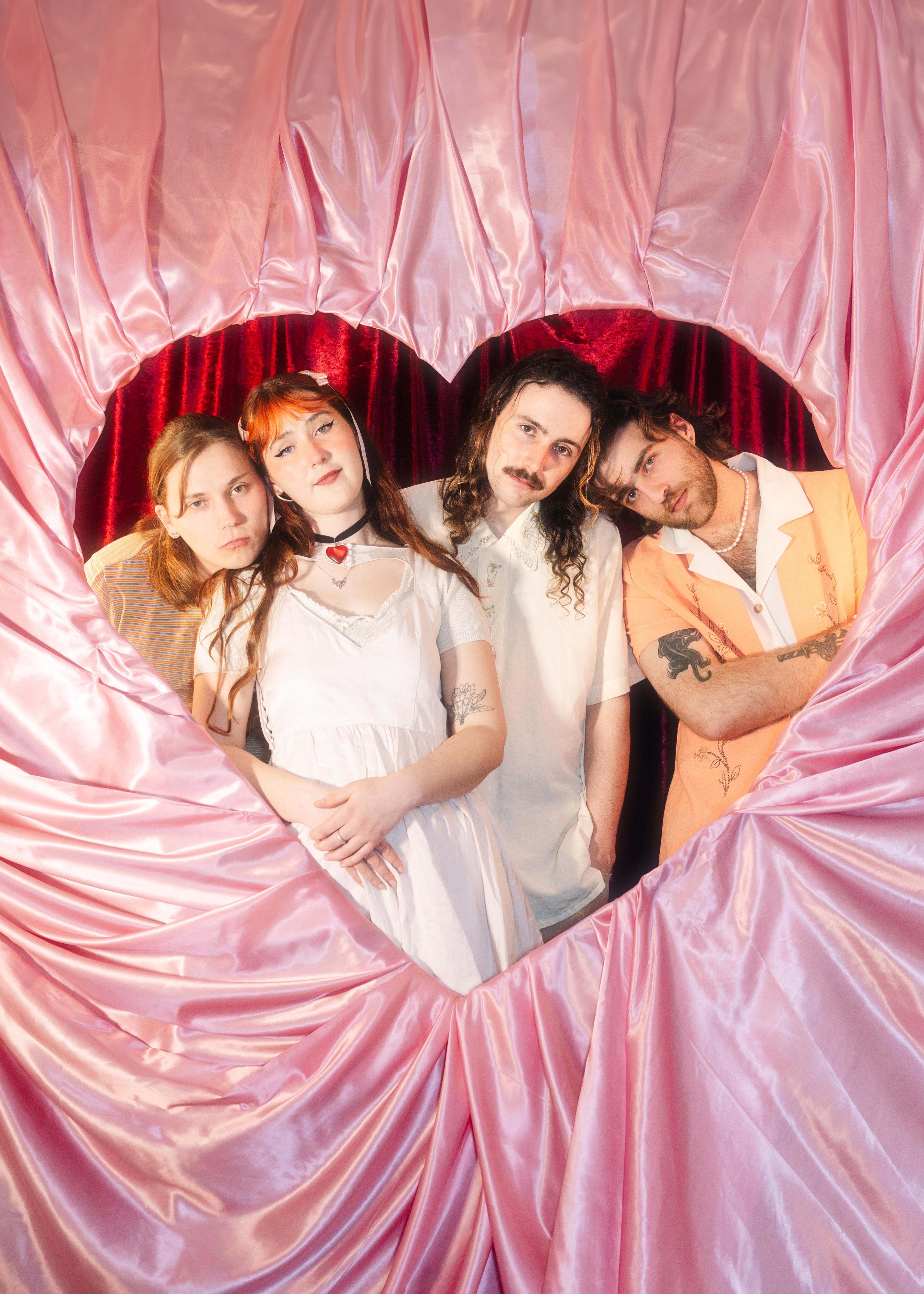 four people stand together in a heart cut out wrapped in pink satin with a red curtain background