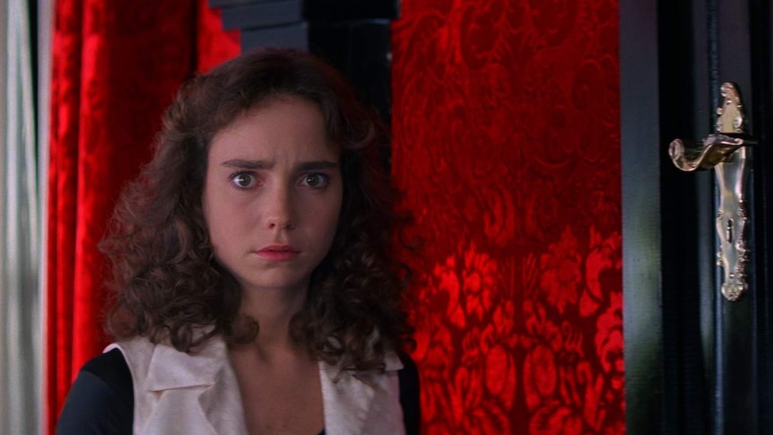 A still from the film Suspiria, with actress Jessica Harper with a concerned expression in front of a vivid red wall.