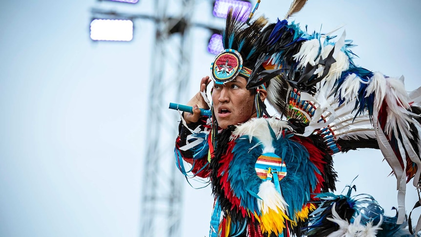 Outdoors on clear day under bright lights a man in vibrant black, blue and white Native American regalia poses with hand to ear.