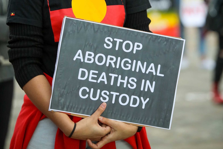 A close-up shot of a sign reading 'Stop Aboriginal deaths in custody' held by a person wearing a shirt with an Aboriginal flag.