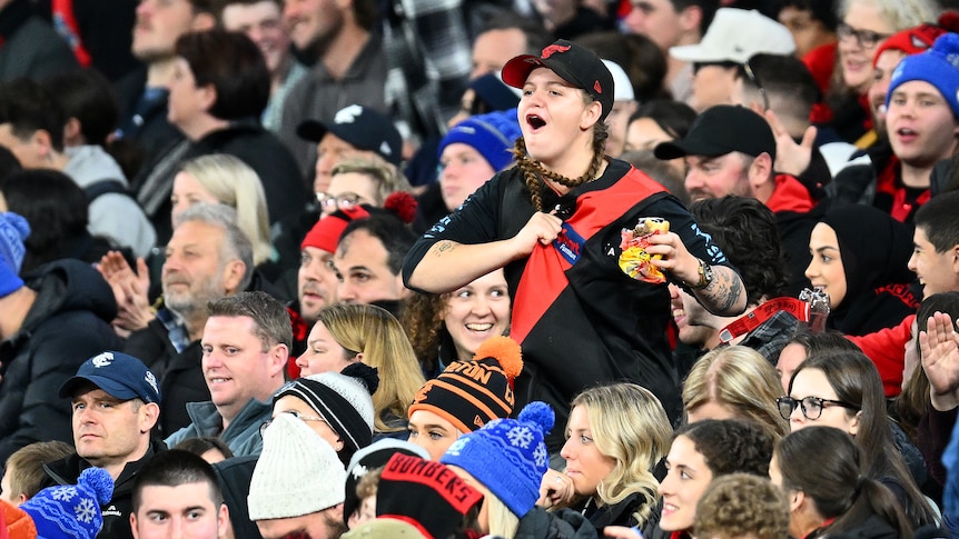 An Essendon fan stands up in the crowd and tugs at her Bombers jumper while cheering.