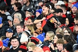 An Essendon fan stands up in the crowd and tugs at her Bombers jumper while cheering.