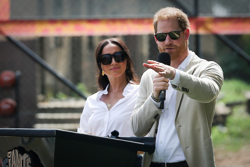 Prince Harry and Meghan wearing white shirts and sunglasses stand infront of a podium.