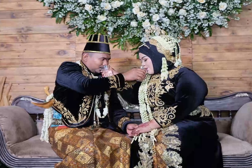 A bride and groom in traditional Javanese garb hold glasses of water to each other's mouths
