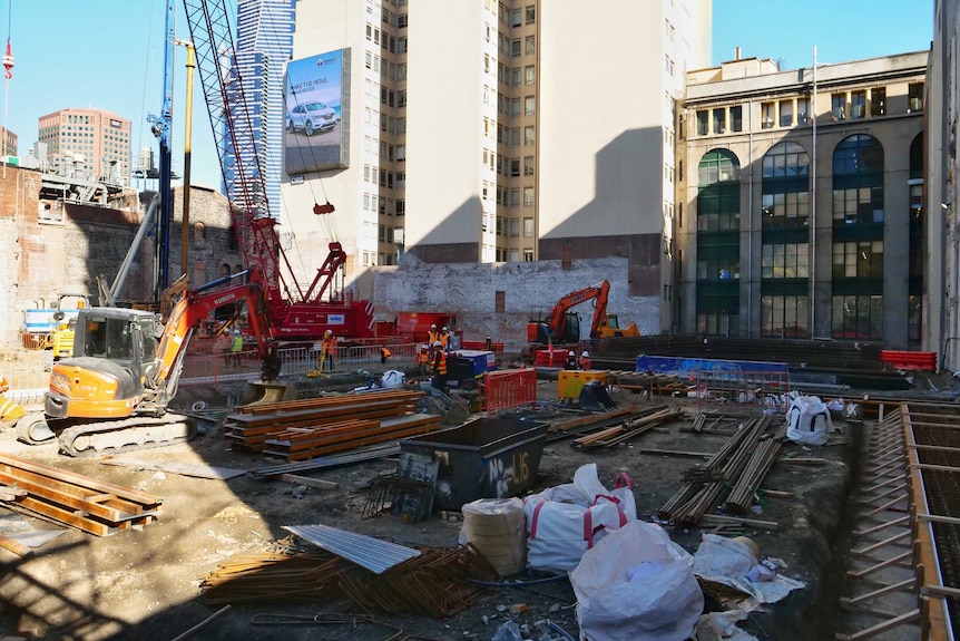 Diggers, cranes and construction workers in a construction site surrounded by tall buildings.