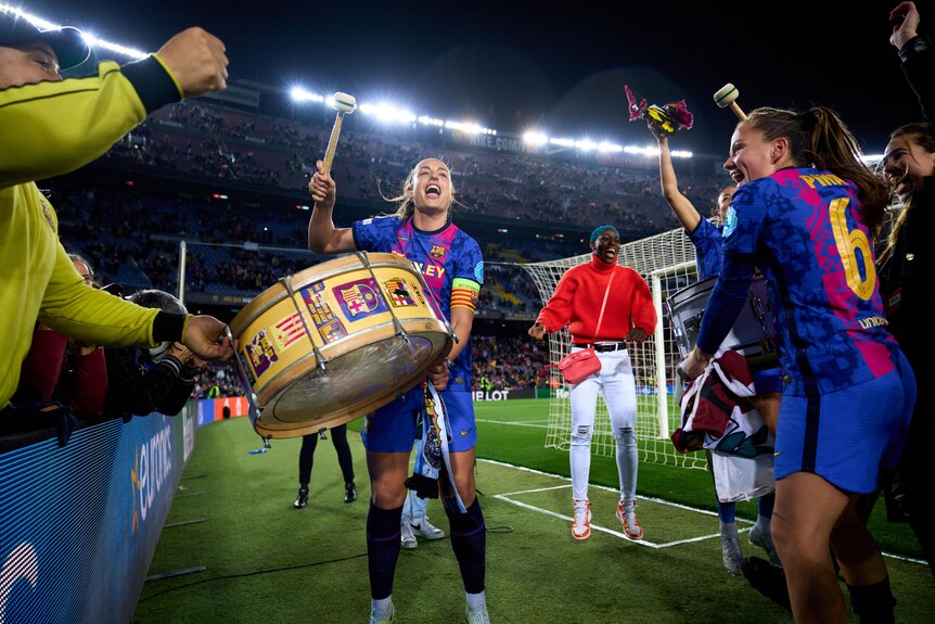 A joyous women's footballer strikes a big drum as her teammates and fans celebrate a win. 