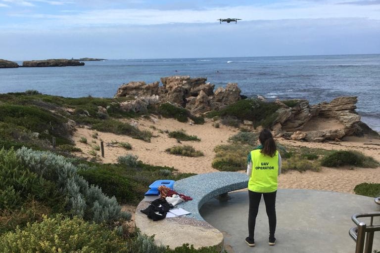 A woman in a yellow vest stands next to a concrete bench with her back to the camera, a drone hovering above at the seaside