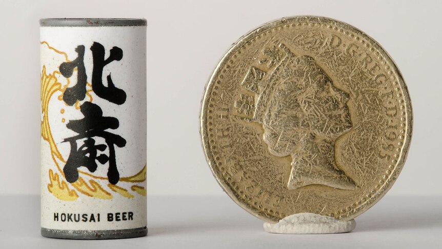 Colour photograph of a miniature Hokusai beer can prop next to a British Pound for scale.