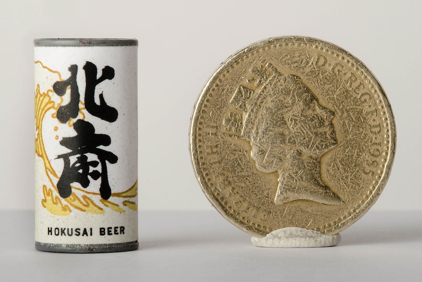 Colour photograph of a miniature Hokusai beer can prop next to a British Pound for scale.