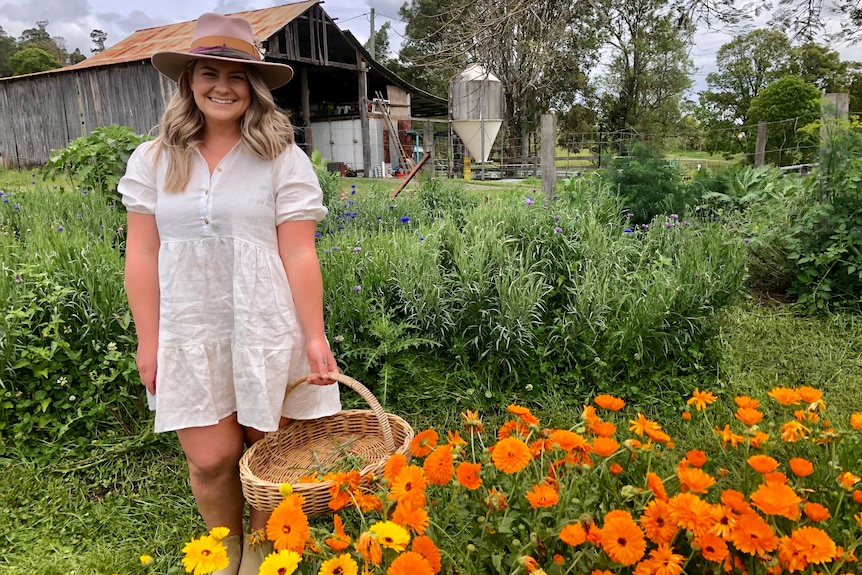 A standing woman smiles behind a row of beautiful orange flowers with a rustic barn in the background.