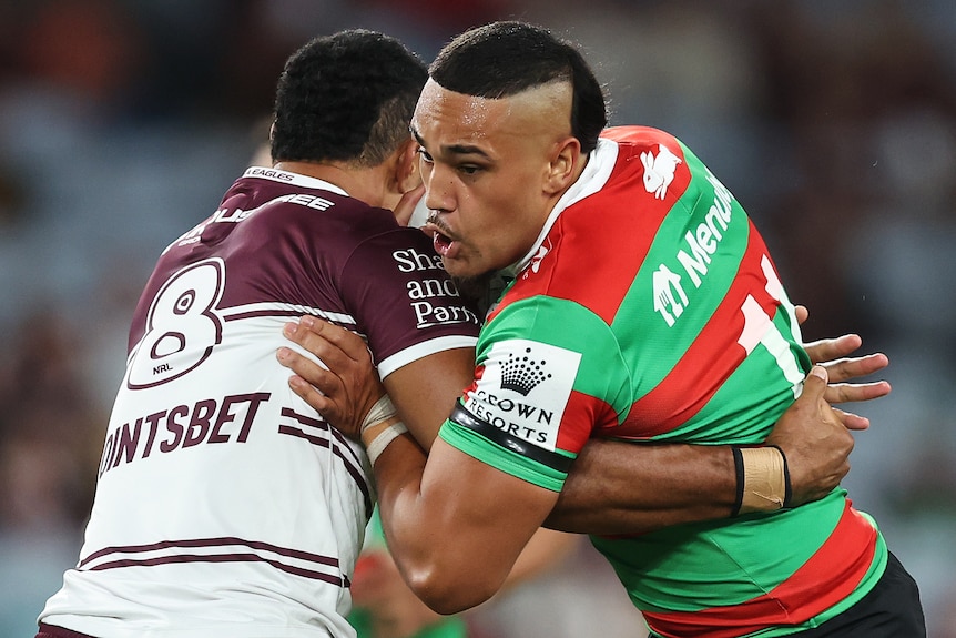 A South Sydney NRL player carries the ball as he is tackled by a Manly opponent.
