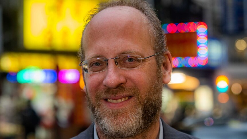 A head shot of a man smiling with bright signs behind him that are out of focus