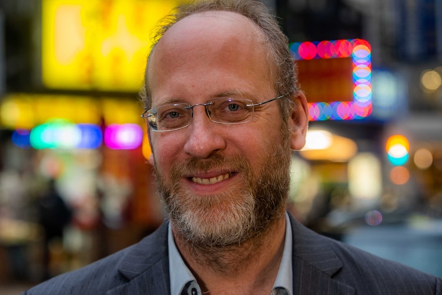 A head shot of a man smiling with bright signs behind him that are out of focus