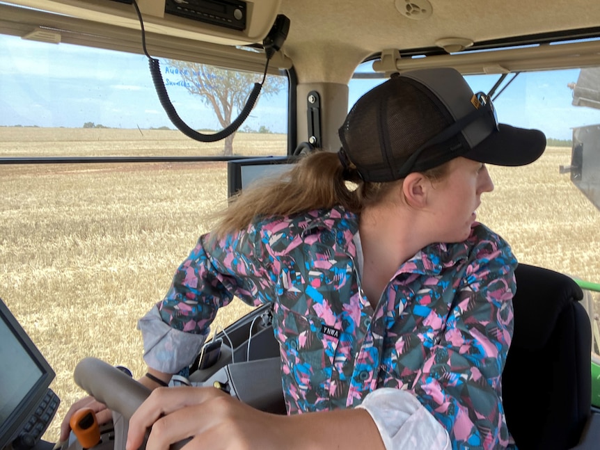 A teenage girl driving a tractor and look at the grain being unloaded into the chaser bin she is towing.