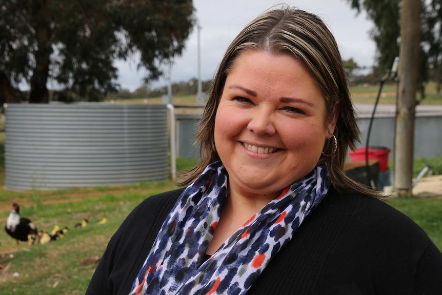 A woman in a black top and coloured scarf stands outside on a farm with chickens and a water tank in the background.