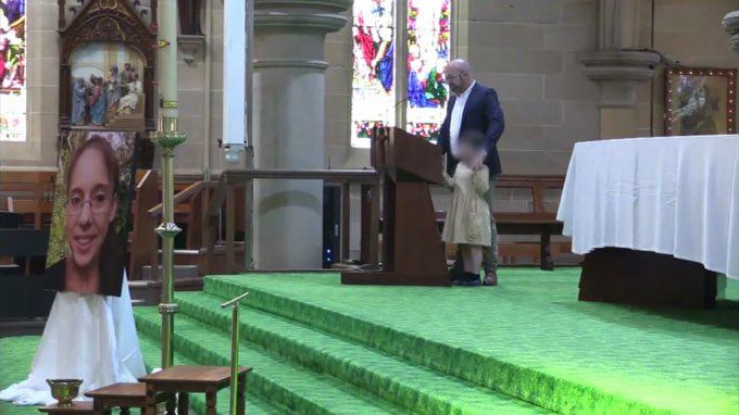 In a church, a man gives a speech from the alter above a woman's coffin and an enlarged photo of her