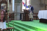 In a church, a man gives a speech from the alter above a woman's coffin and an enlarged photo of her