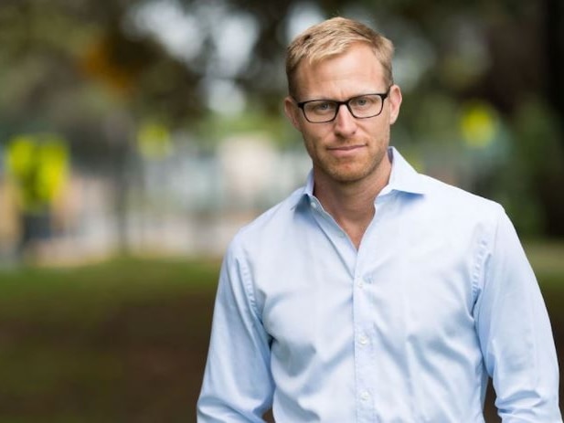 A slim, blond, bespectacled man standing in a leafy area.