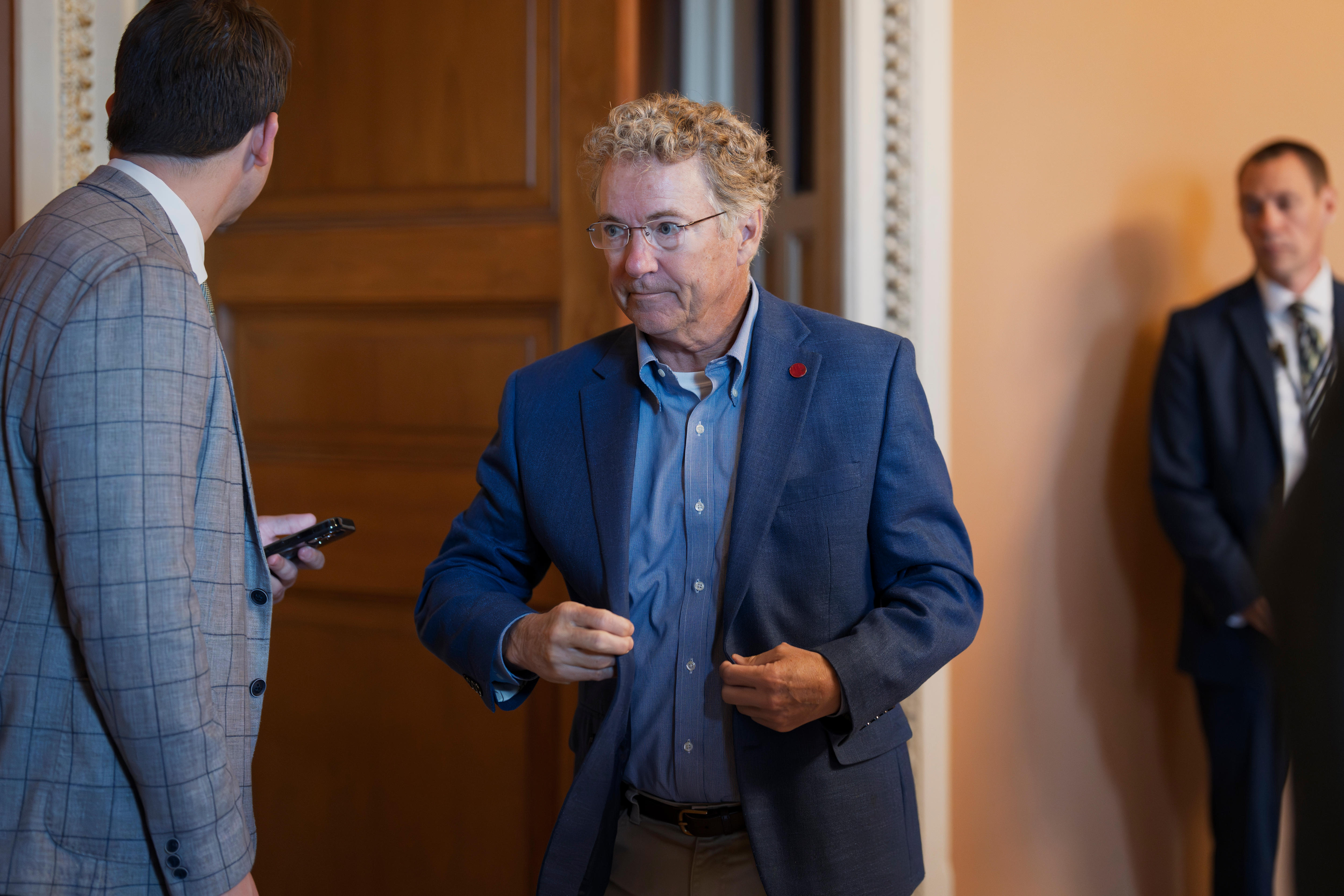 A curly-haired man with a navy blazer and light blue shirt walks out of a room with a large, wooden door